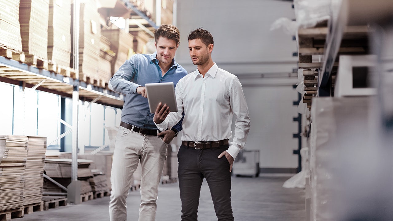 Two men wearing business casual clothing discuss information on tablet device. 