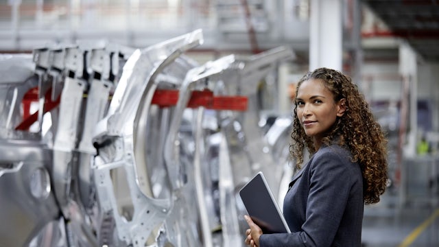 A woman wearing a business jacket and holding a tablet device stands in automotive factory line. 