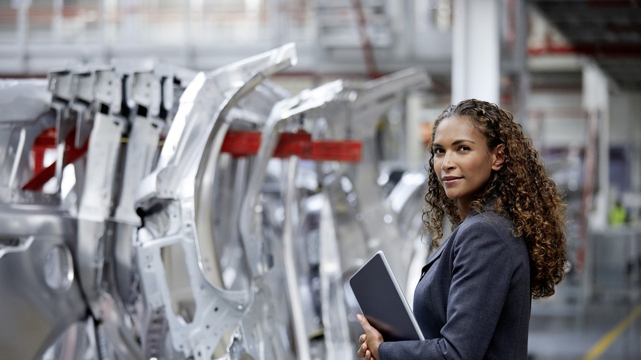A woman wearing a business jacket and holding a tablet device stands in automotive factory line. 