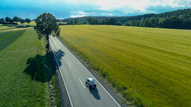 A car driving down a 2-lane road in the middle of grassy fields and trees.