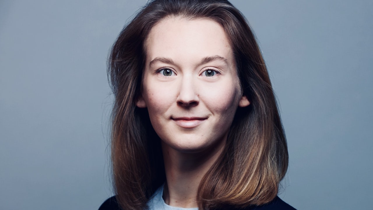 A woman with short hair and a serious expression is wearing a blue shirt and has a mole on her right cheek.