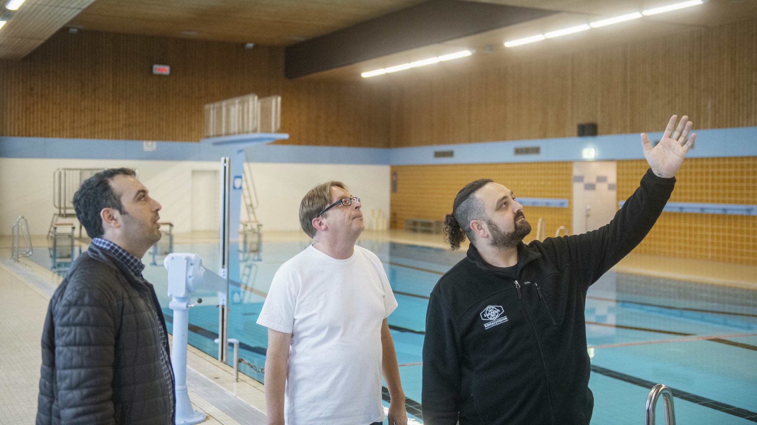 Three men look upward while standing on the deck of an indoor pool.