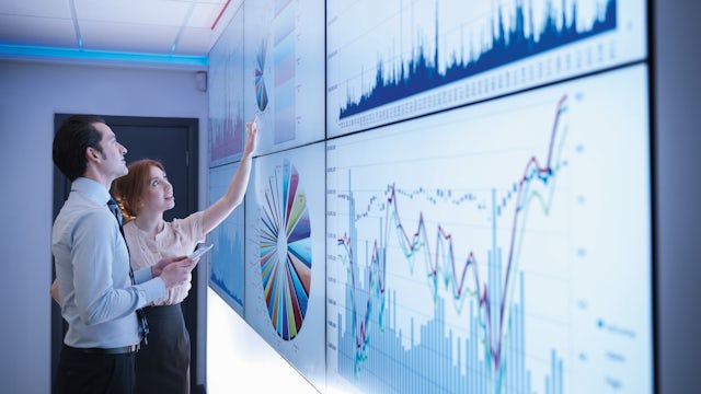 A stack of hundred dollar bills with a calculator and pen on top, sitting on financial documents and graphs.