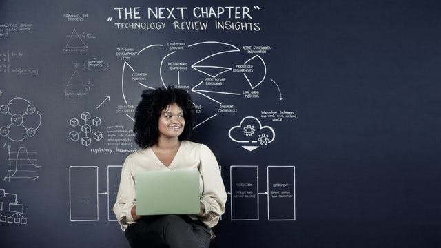 Young woman on a stool with an open laptop in front of a chalkboard.