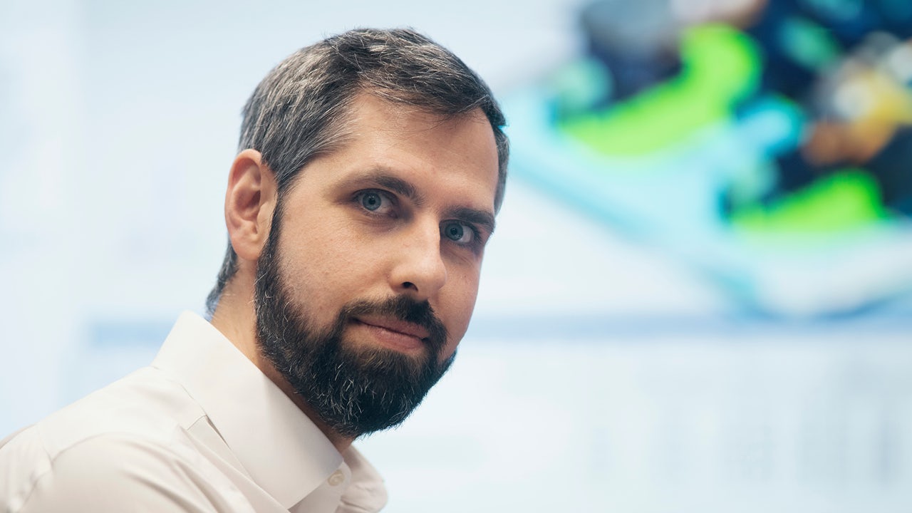 Person in black shirt standing against white wall, holding a dark object with blurred background.