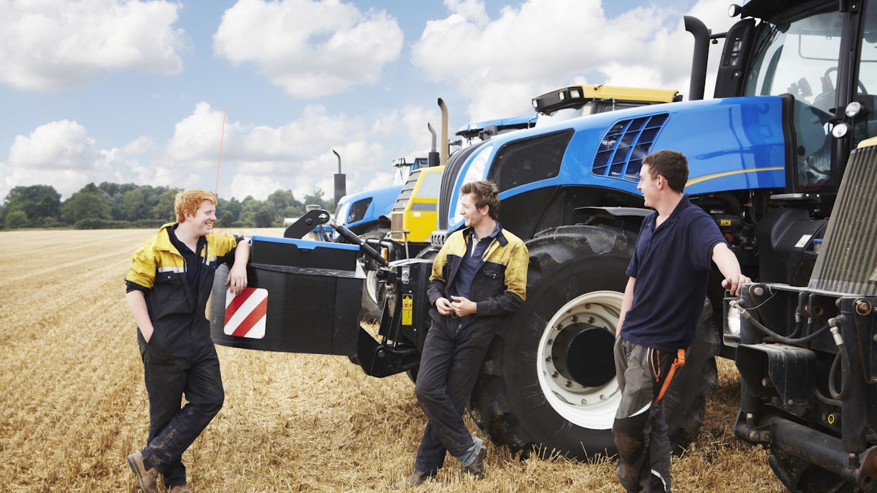 Two farmers are standing near a machinery vehicle in a field, talking to each other.