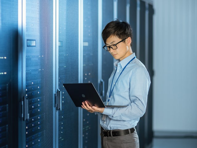 A stands in front of a large bank of computer servers while he works on a laptop.