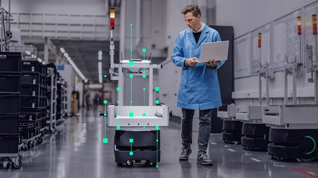A man in a lab coat uses a laptop next to an automated guided vehicle (AGV) with green data overlays in a factory setting.