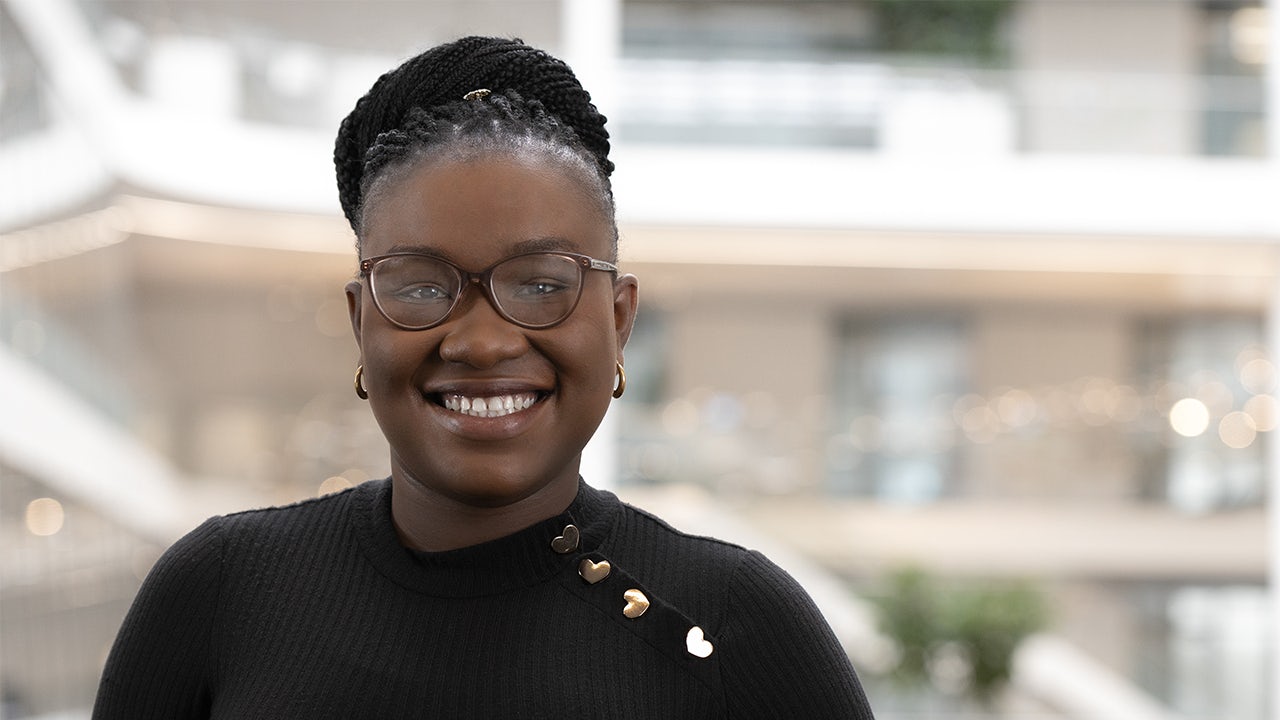 A woman in a white shirt stands in front of a Siemens building, smiling and looking at the camera.