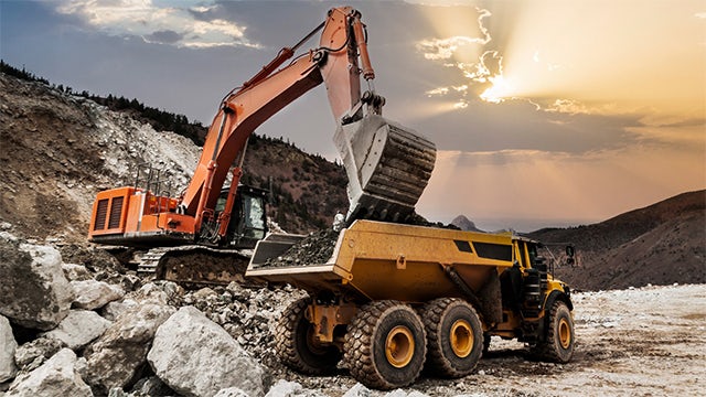 An excavator and a dump truck moving rocks near a mountainside.