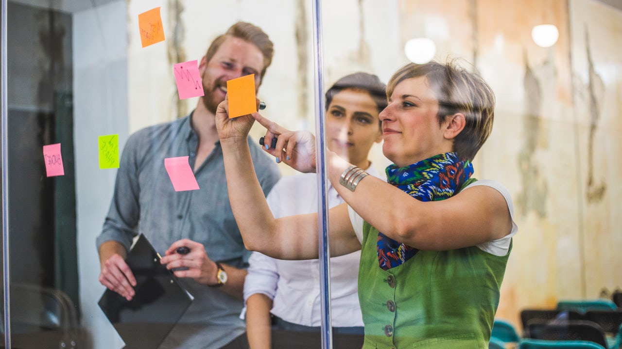 A person is standing in front of a white wall with a blue and white logo on it.