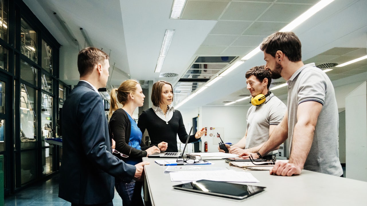 A person is standing in front of a white wall with a blue and white logo on it.