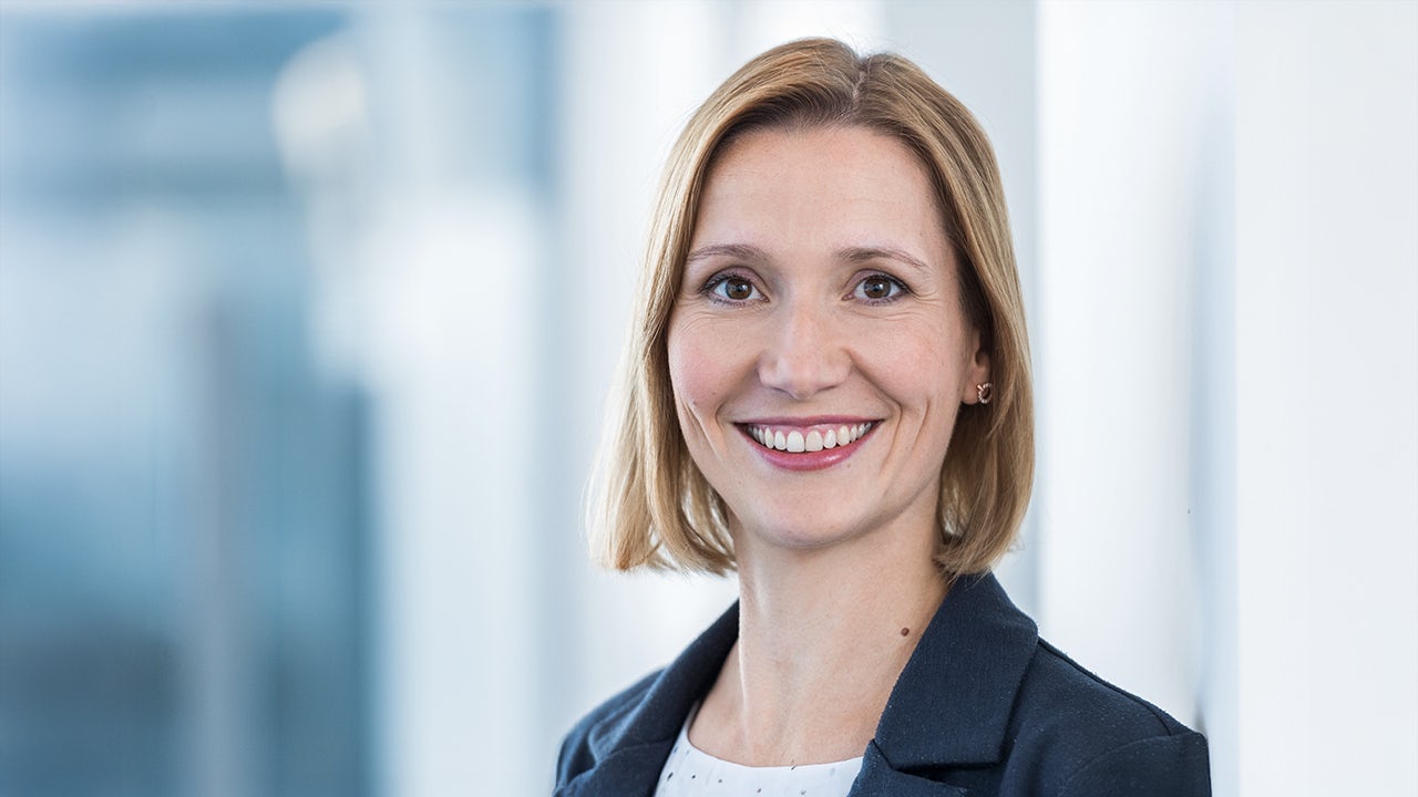 A woman with short hair is standing in front of a white wall, wearing a black top and looking directly at the camera.