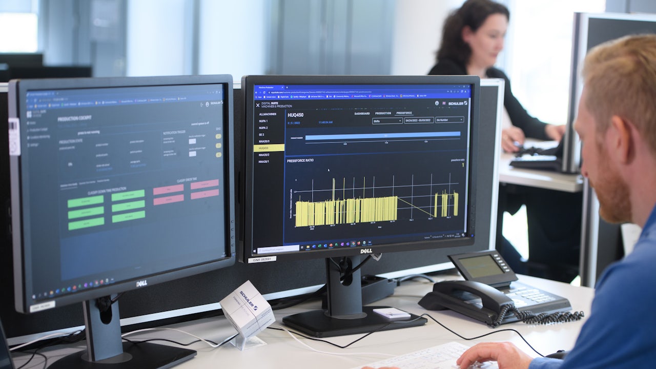 A man sits at a desk with two monitors displaying production data.