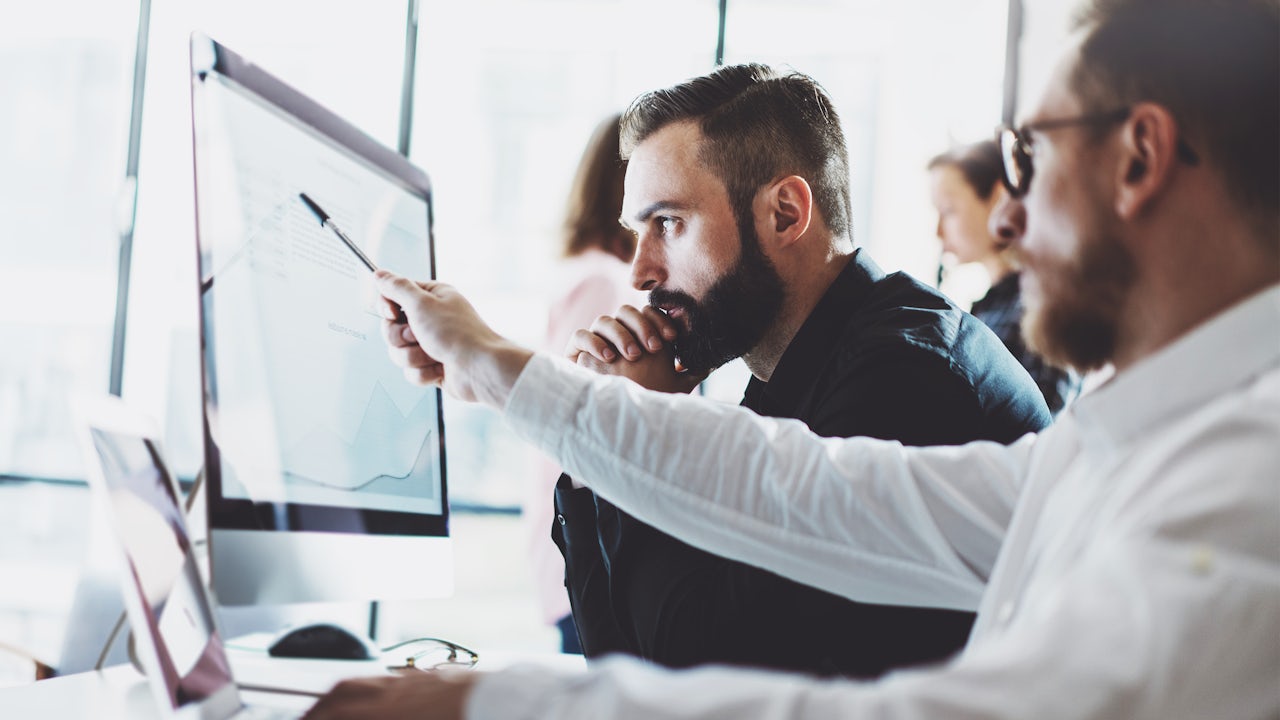 Two men sitting at a desk, looking at information on a laptop and a computer monitor.