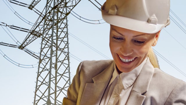 Woman in hard hat and suit smiling while using tablet near electrical transmission tower.