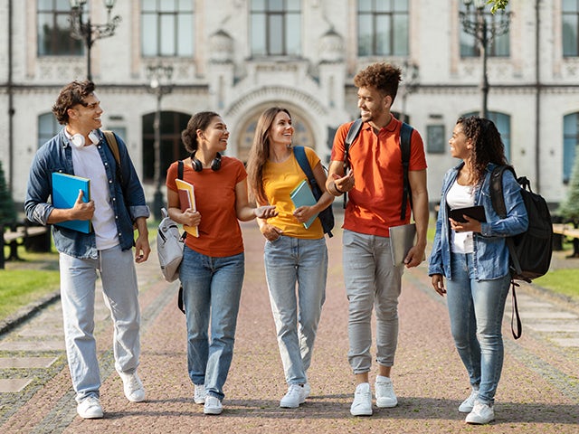 Group of five college kids walking on campus with university building behind them.