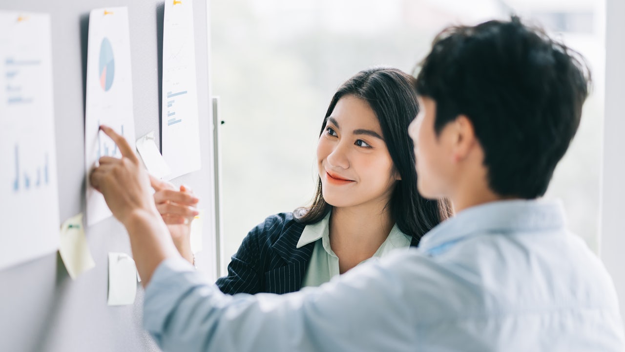 A hand holding a magnifying glass over financial documents with charts and graphs on a wooden desk.
