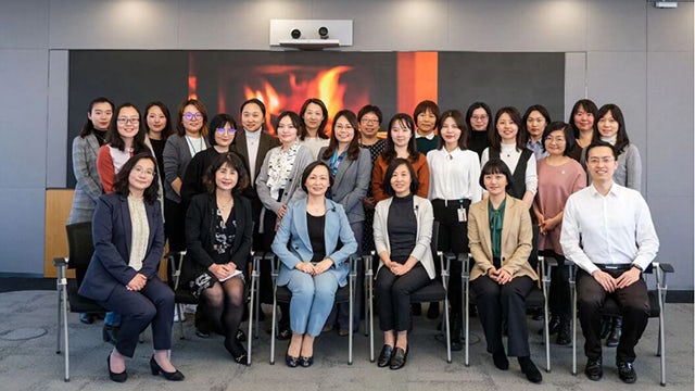 Three smiling women in professional attire standing together in an office setting with computers visible in background