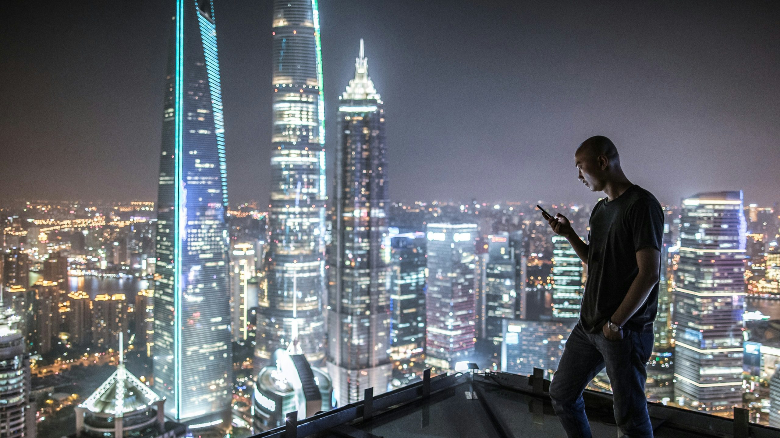 Man in an office overlooking a cityscape of skyscrapers looks at his phone.