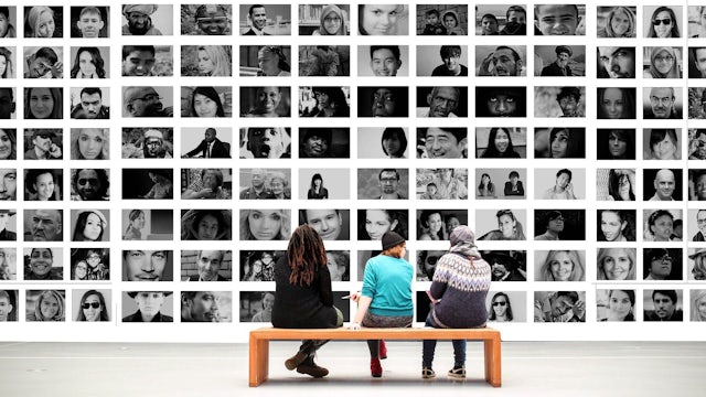 Three people sitting on a bench in front of a wall full of black and white photos of people's faces.