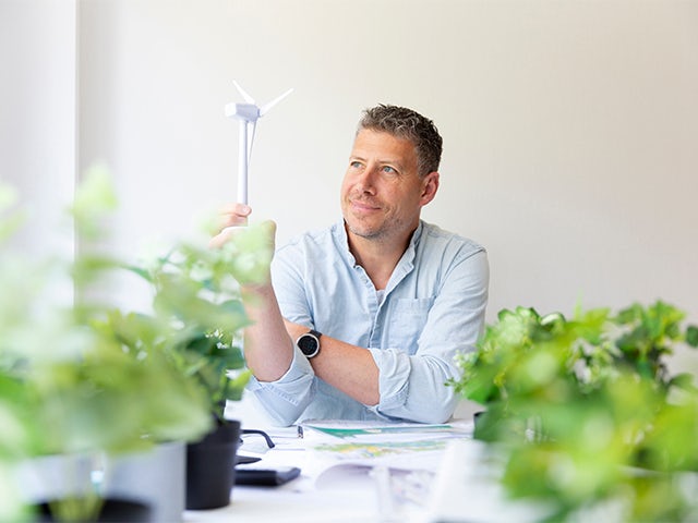 Man sitting at his work table holding a model of a wind turbine in his hands and is surrounded by plants.