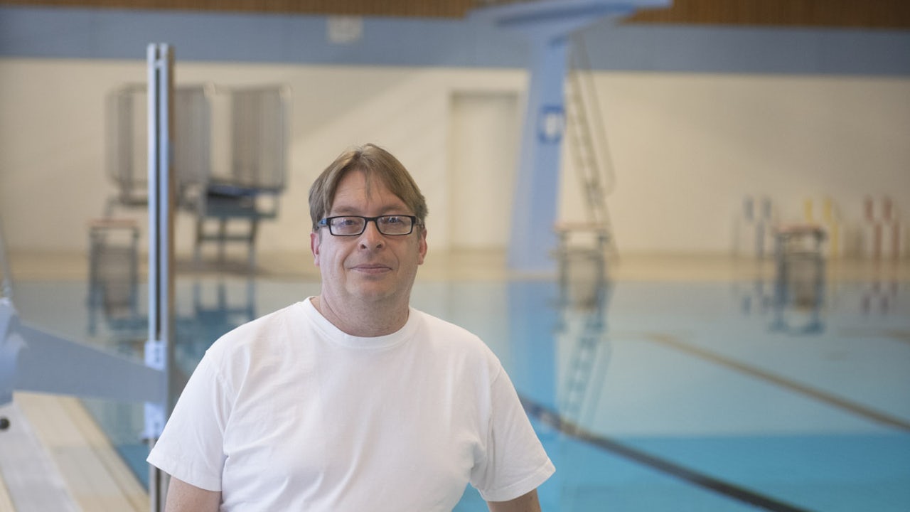 A man in a white t-shirt and glasses stands next to an indoor pool.