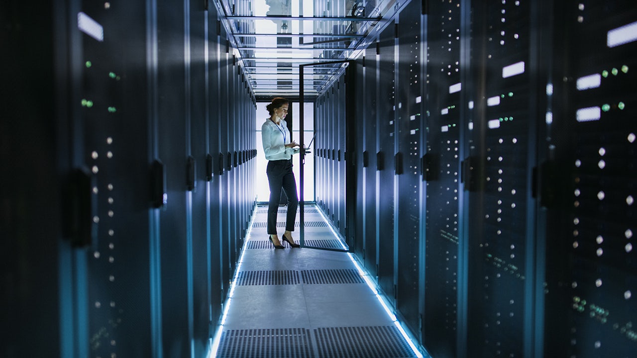 Woman standing in data center, holding a laptop.
