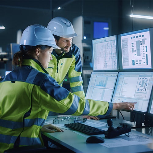 Two engineers in a control room wearing safety gear reviewing data and diagrams on multiple monitors.