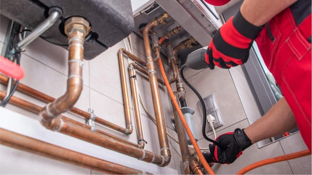 A person wearing red safety gloves and using a digital tool to perform an inspection on a pipe connection.