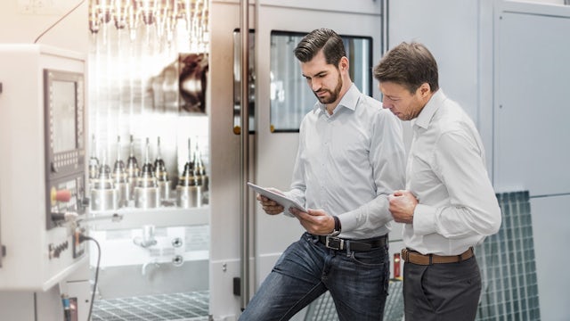 A person in a suit is standing in front of a desk with a computer monitor, a cup, and a book, talking on the phone.