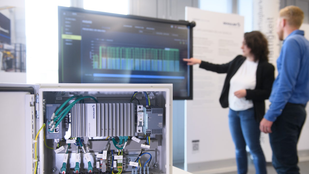 Foreground close-up of hardware in a cabinet with two blurred people and a screen in the background