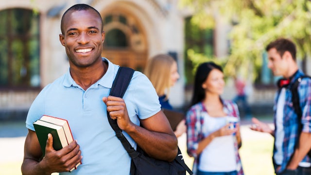 A college student, wearing a backpack, holding a book, and smiling, while his classmates are in the background talking.