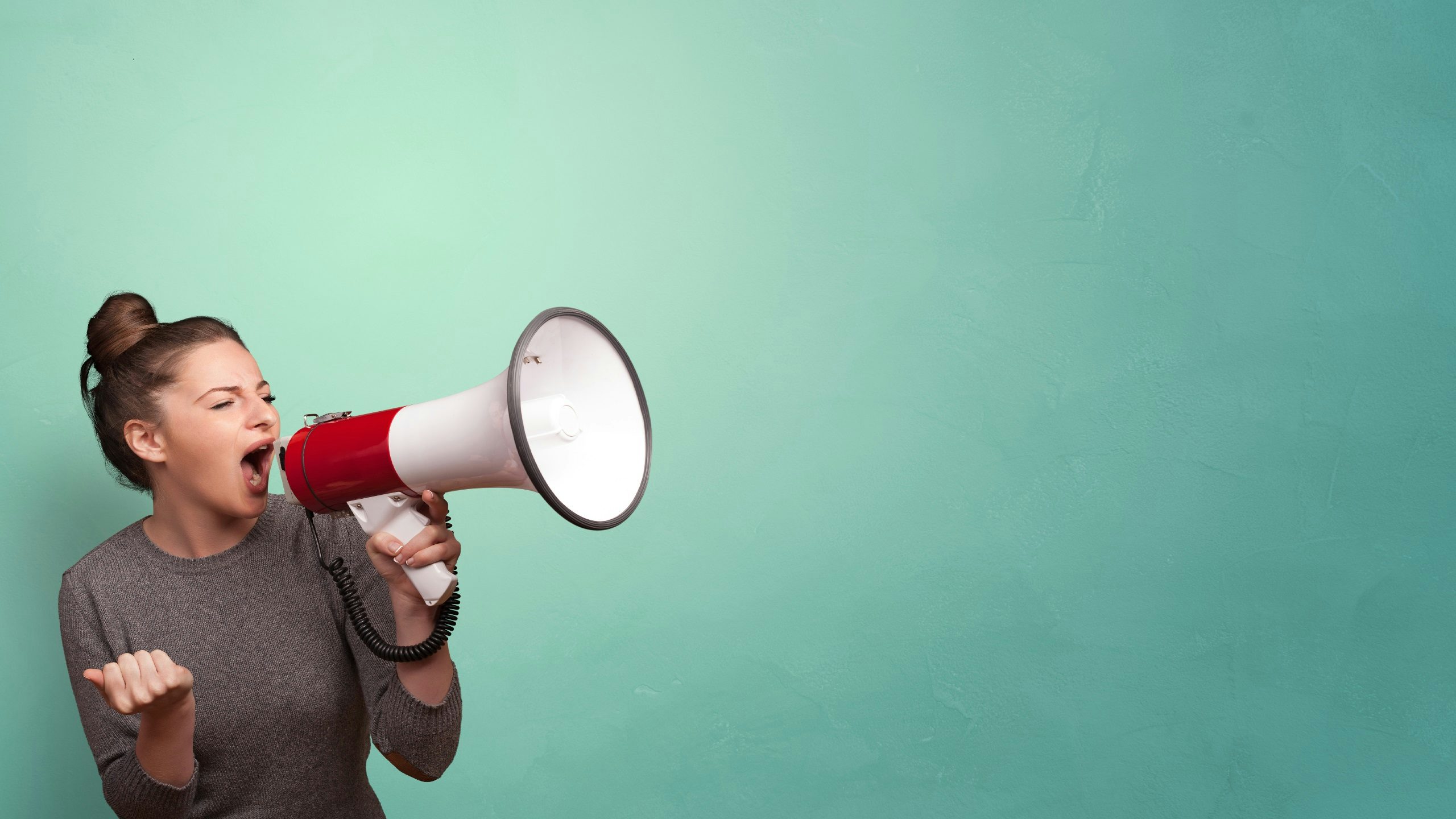 A woman wearing a grey long sleeve shirt shouting into a megaphone against a blue background.