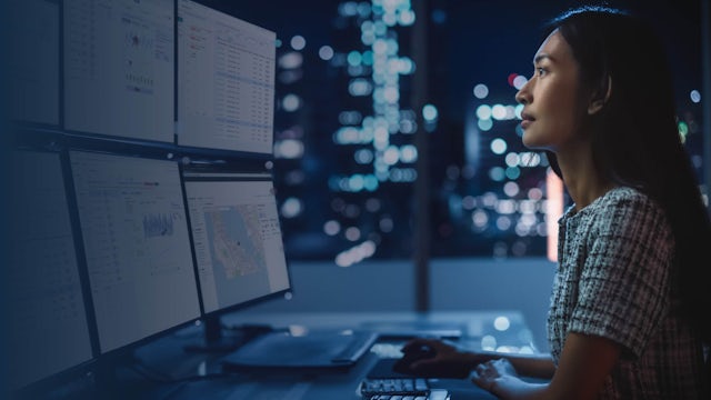 Woman looks at multiple monitors in a darkened office in front of a window with lit buildings in the background