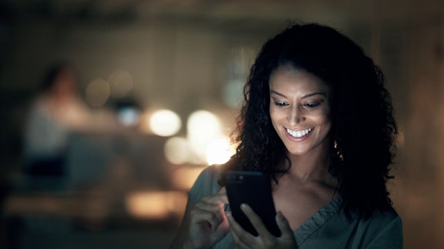Woman using mobile phone at night with warm lights in background.