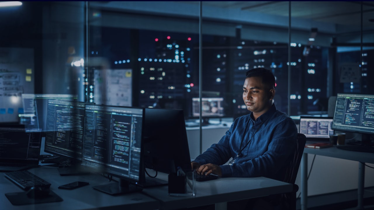 A man uses Gridscale X software on his computer at night, with lighted buildings in the background.