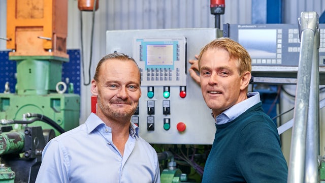 2 men standing in front of a machine in a factory, staring and smiling at the camera.