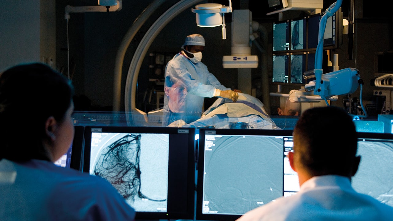 A view from behind of two people watching computer monitors while a surgeon works on a patient in the background.