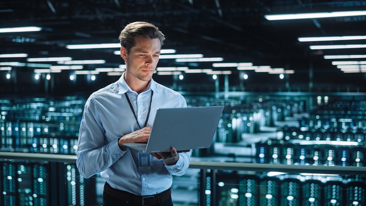 Employee stands in warehouse and reviews open laptop.