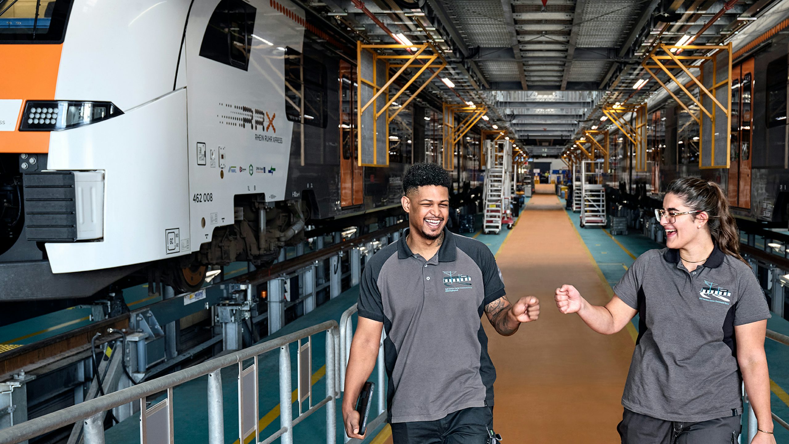 Two rail technicians walking and talking inside a train maintenance facility with trains on either side.