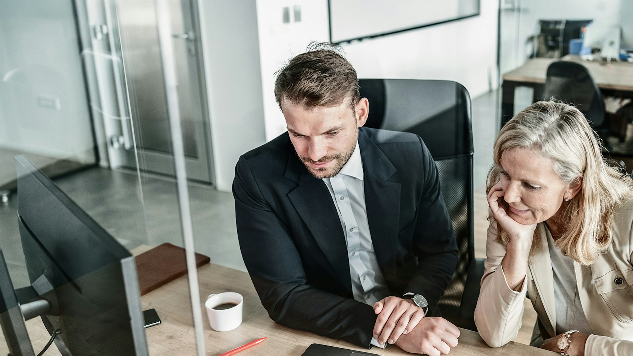 Two colleagues of different generations reviewing information together on a computer at a desk in a modern office.
