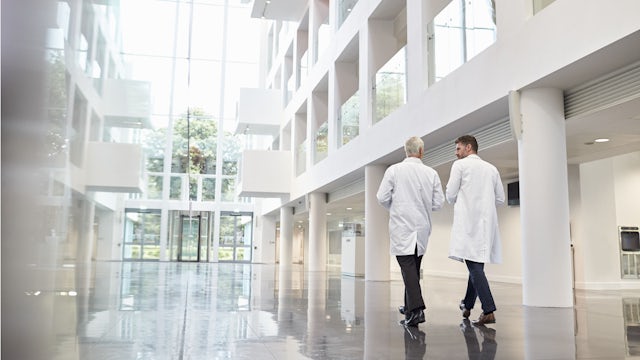 Two doctors in lab coats walk down a hallway in a medical facility. 
