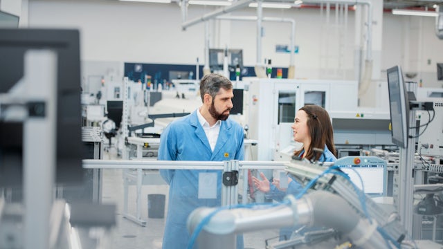 Two coworkers chat behind a manufacturing cell on a shop floor.
