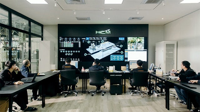 HCM employees gather in a conference room and look up at a large screen.