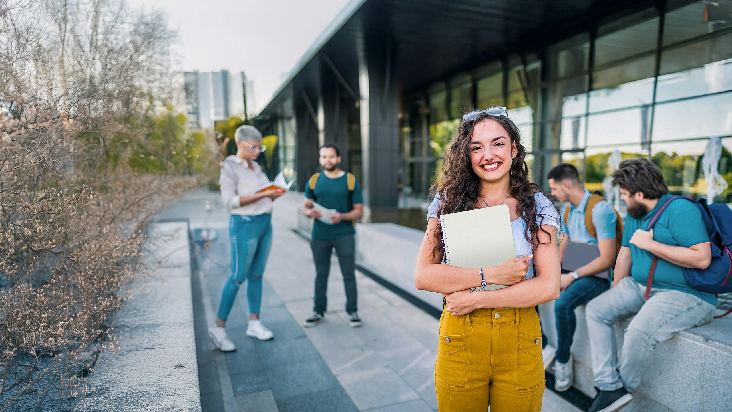 Young woman with curly hair smiling while holding books and a laptop on a college campus
