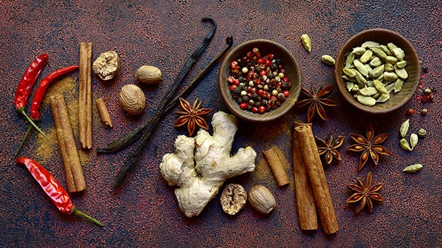 Assortment of spices arranged on a textured surface, including cinnamon sticks, dried chilies, ginger root, star anise, nutmeg, vanilla beans, cardamom pods, and bowls of mixed peppercorns.