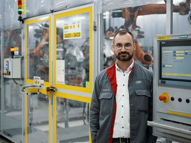 Person standing in an industrial production area next to a control panel, with robotic machinery operating behind safety glass in a manufacturing environment.