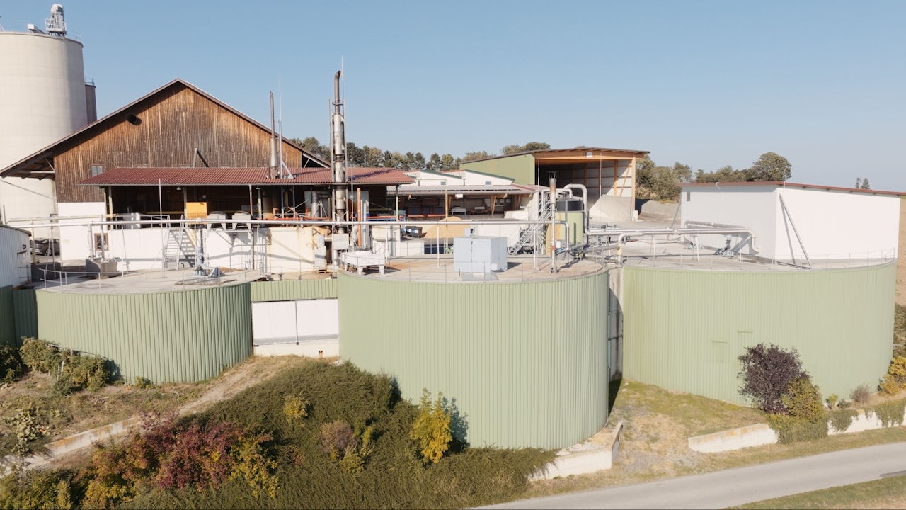 Industrial biogas facility with large green storage tanks, piping, and processing equipment in a rural setting.