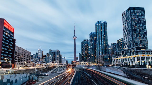 Toronto cityscape at dusk featuring modern high-rise buildings, with railway tracks leading towards the skyline.  
